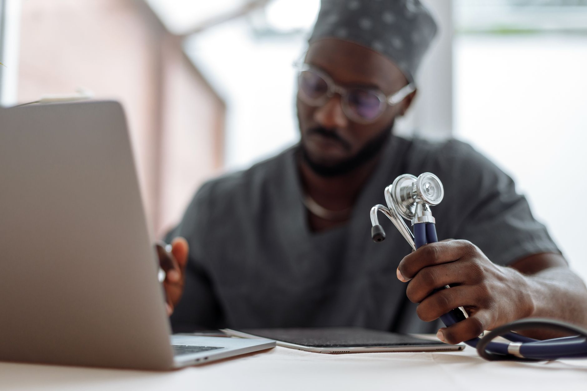 a man in gray scrub suit holding stethoscope while typing on his laptop